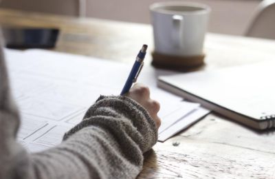 A person writing notes on a printed sheet of paper at a wooden desk beside a mug.