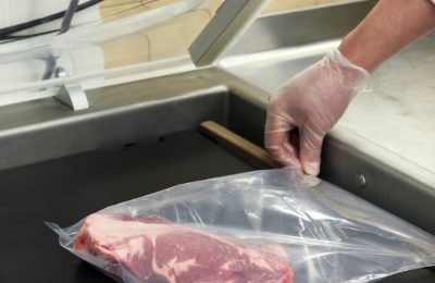 A gloved hand placing a cut of beef into a vacuum pack on a stainless steel bench.